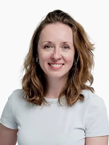 A smiling woman with shoulder-length curly hair wearing a white T-shirt against a pale background.