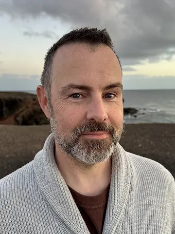 A short-haired bearded man wearing a knitted top on a windswept clifftop by the sea under a cloudy sky.